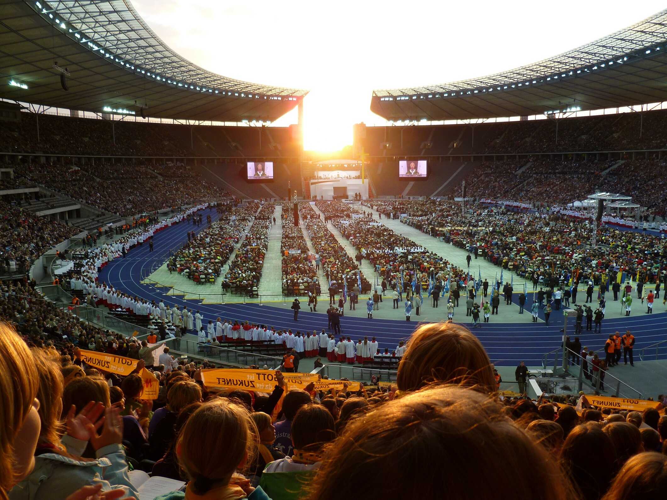 Blick auf volles Stadion, in dem ein Gottesdienst mit Papst Benedikt XVI. stattfindet, im Hintergrund die untergehende Sonne