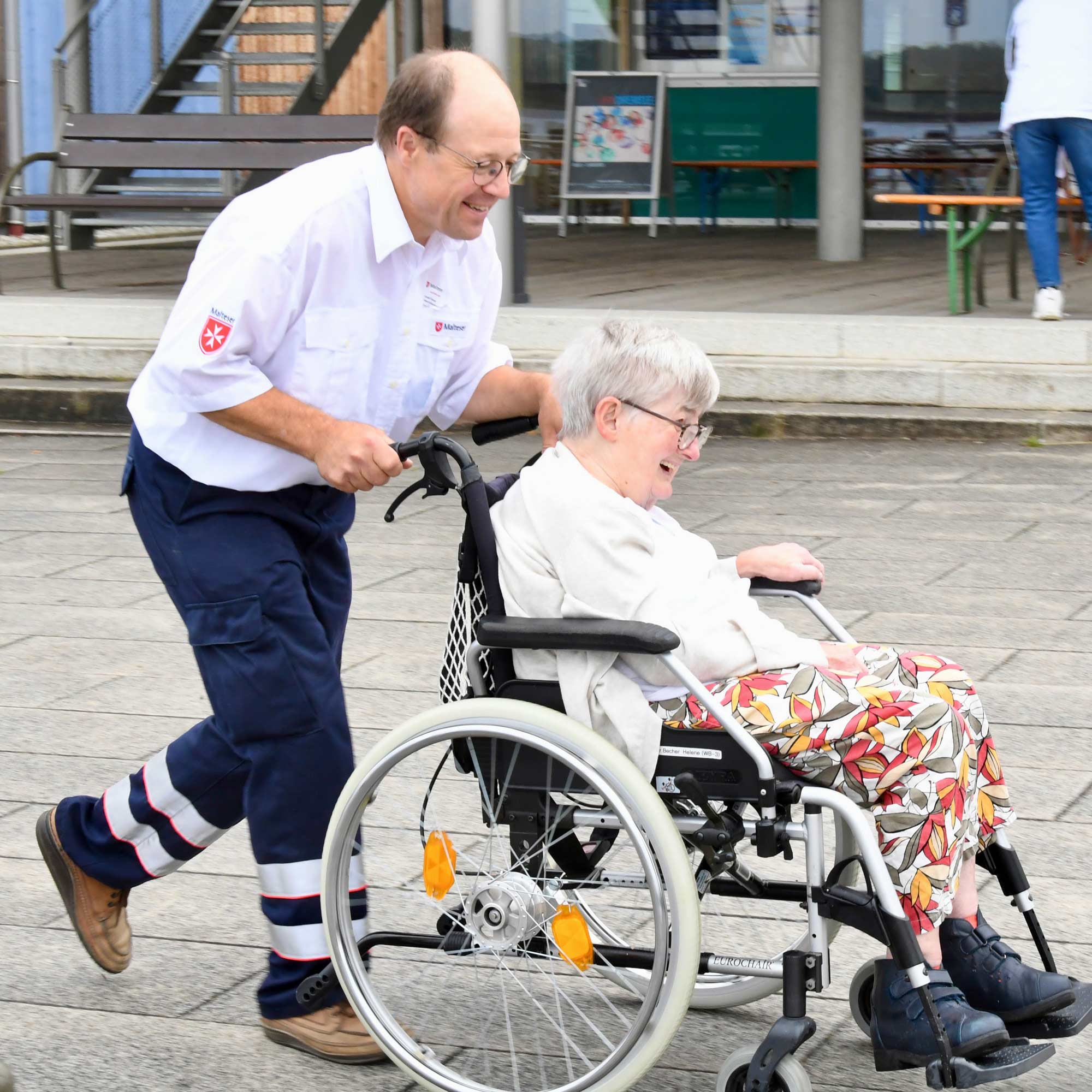 Mitarbeiter eines Hilfsdienstes schiebt alte Frau im Rollstuhl über Bahnsteig