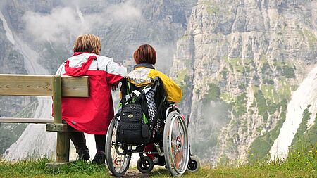 Behinderte Frau im Rollstuhl neben Frau auf Bank mit Blick auf massives Gebirge