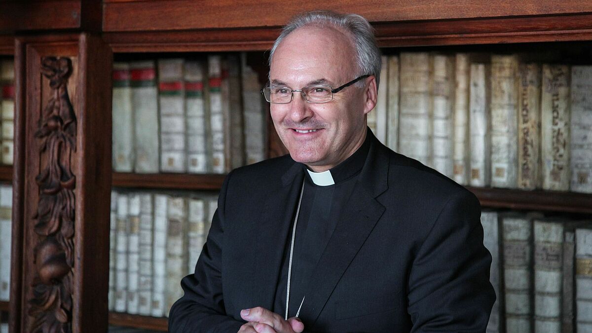 Bischof Voderholzer faltet die Hände vor einem Bücherregal