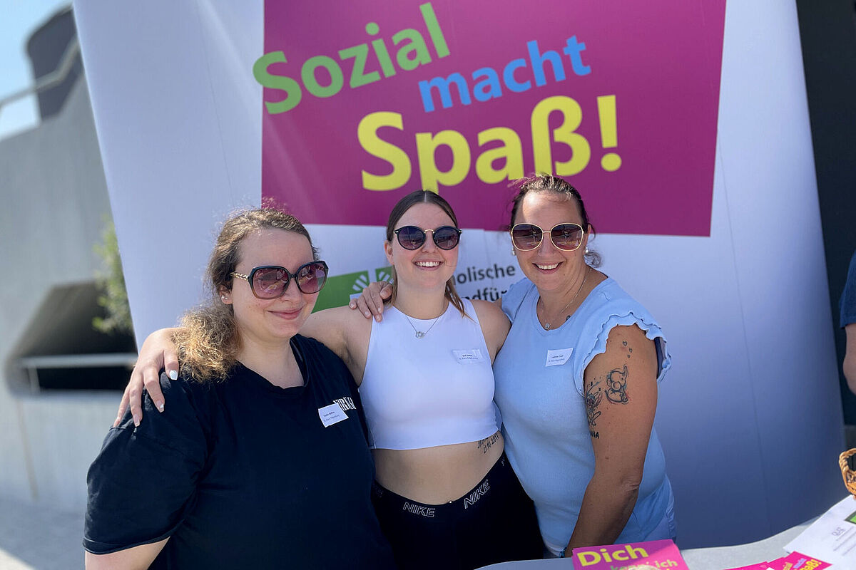 Drei junge lachende Frauen mit Sonnenbrillen, im Arm haltend vor einem Plakat, das für soziale Berufe wirbt