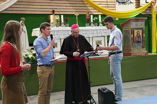 Bischof zwischen zwei jungen Männern mit Mikrofon in der Hand