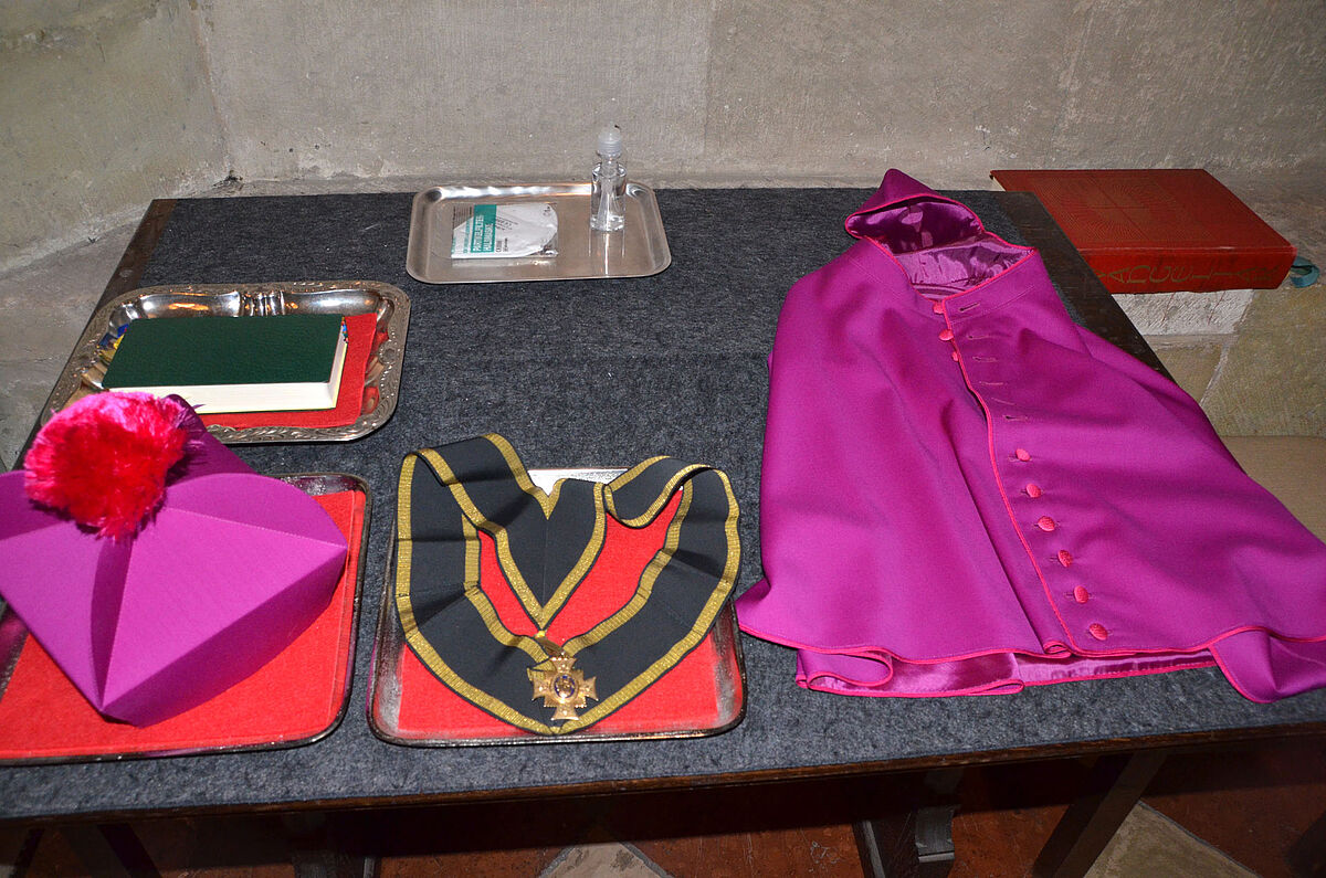 Brevier (Stundenbuch), Birett (priesterliche Kopfbedeckung), Kapitelkreuz und Mozetta (violetter Schulterkragen) liegen auf einem Tisch aus.